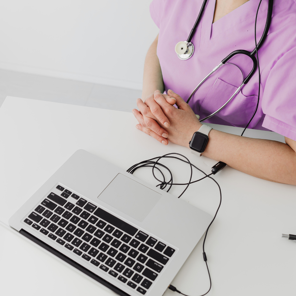doctor in scrubs sits at a table with an open laptop. she is wearing a smart watch and has a headset connected to the PC